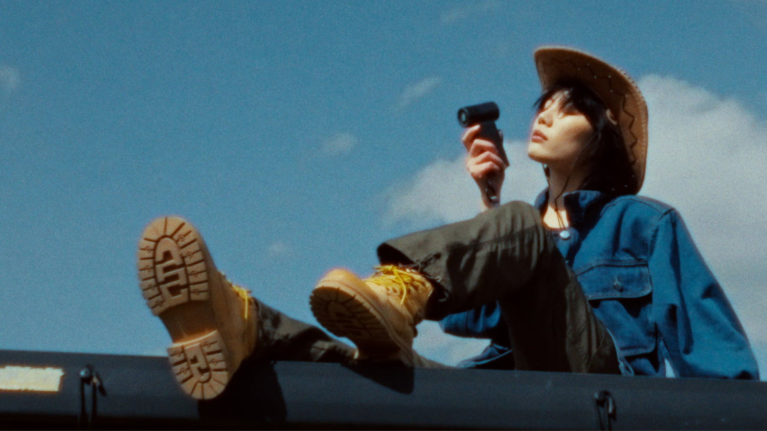 Woman wearing a cowboy hat sits outdoors holding a compact handheld fan against a blue sky.