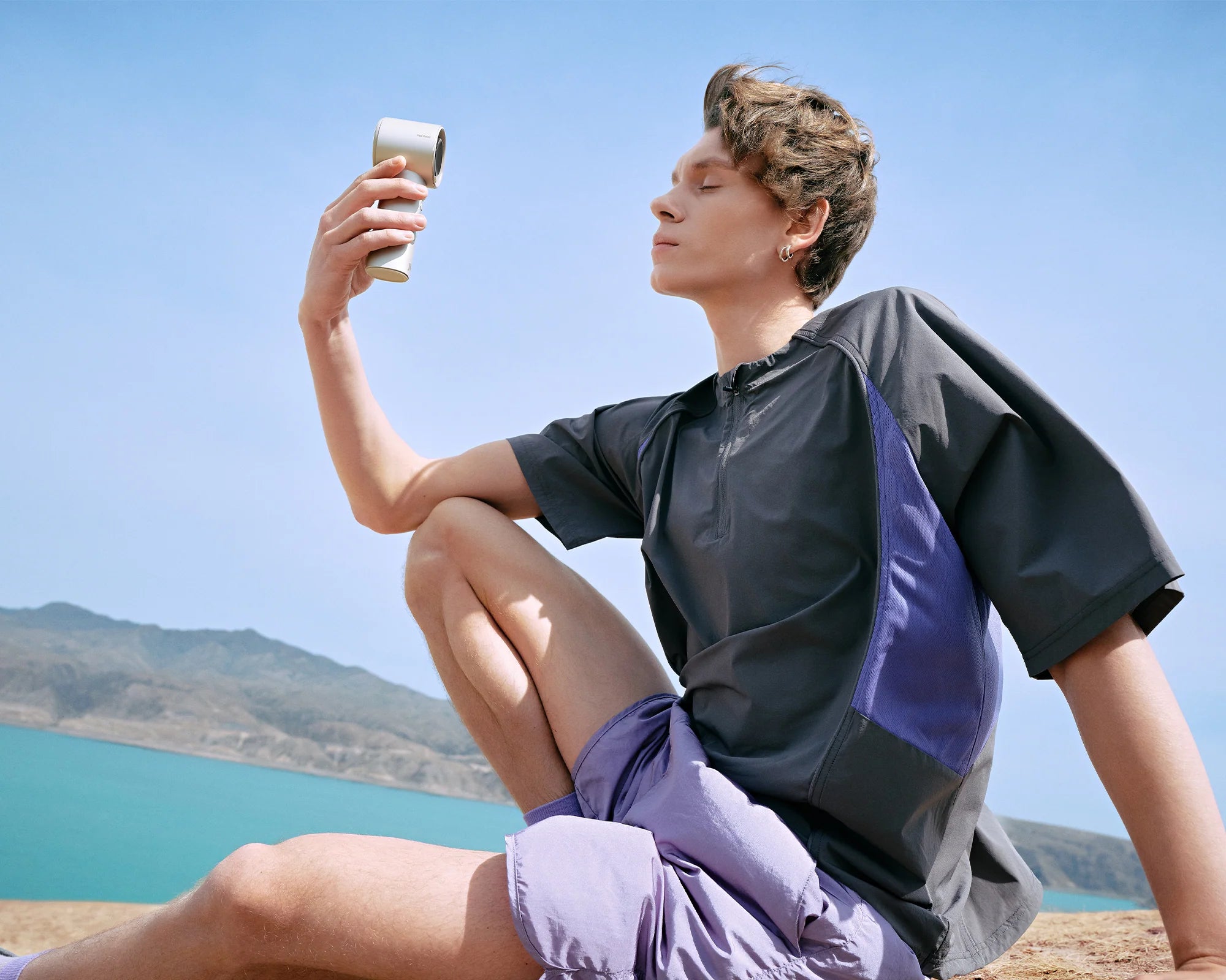 Man uses a portable handheld fan while resting by a sunny mountain lake